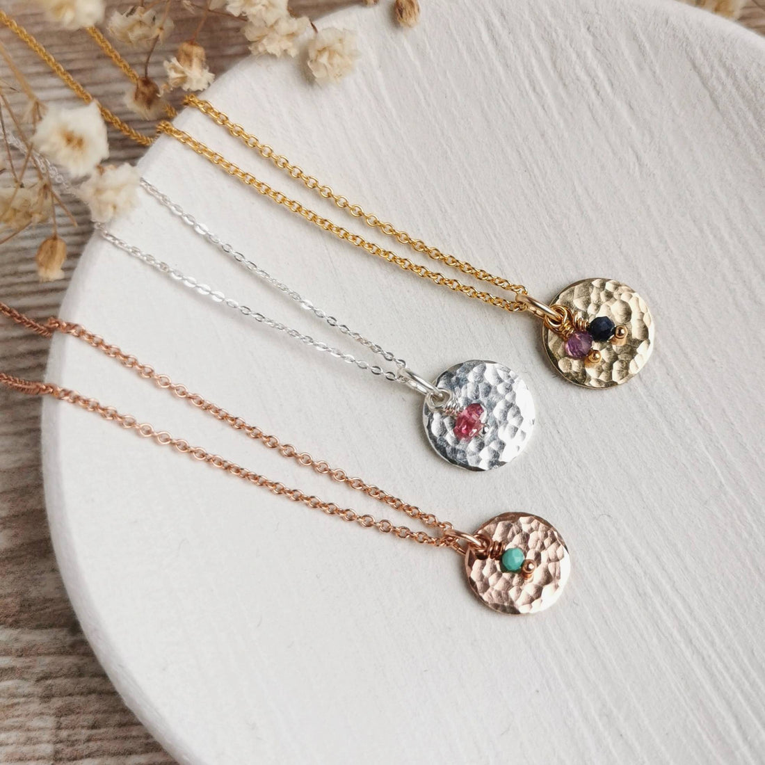 gold, silver and rose gold necklaces with birthstone charms laid on a ceramic dish with dried flowers