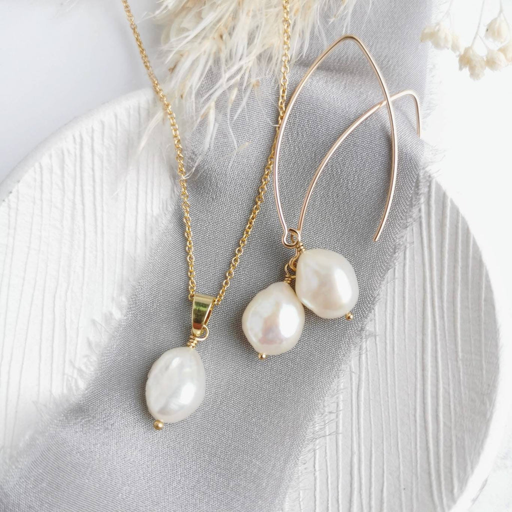 A set of gold and ivory baroque pearl earrings and a matching necklace laid out on a white surface, with the pearl earrings hanging from 14 carat gold-filled hooks and the necklace with a 14k gold-filled chain.