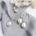 A set of ivory baroque pearl earrings and a matching necklace laid out on a white and grey surface, with the pearl earrings hanging from silver hooks and the necklace with a silver chain