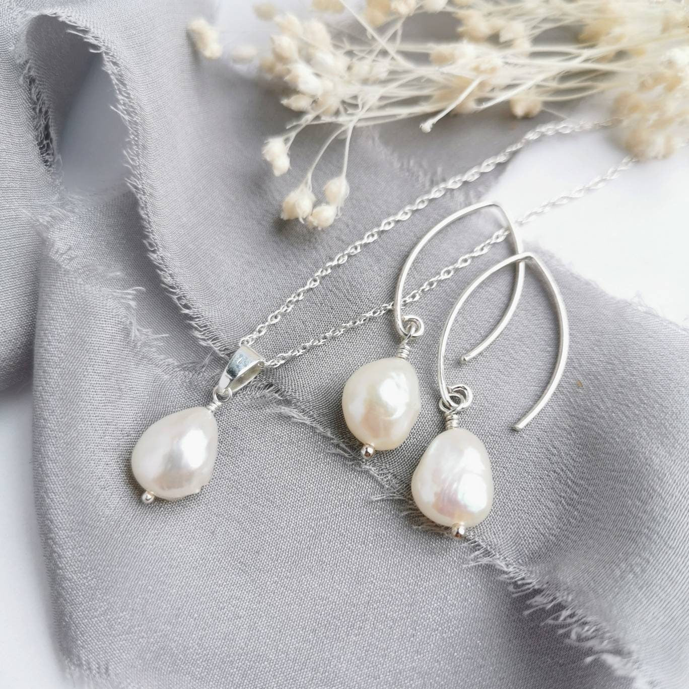 A set of ivory baroque pearl earrings and a matching necklace laid out on a white and grey surface, with the pearl earrings hanging from silver hooks and the necklace with a silver chain