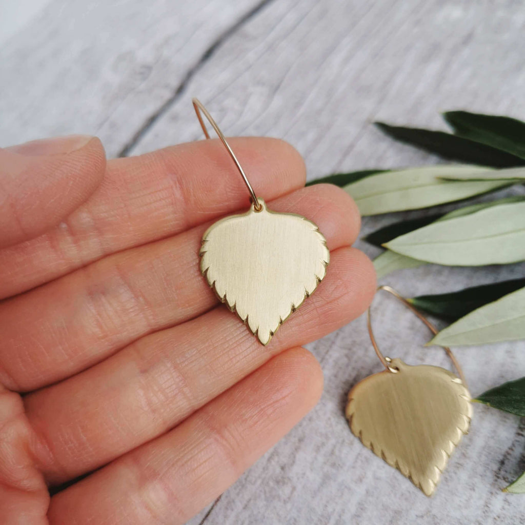 Golden brass birch leaf-shaped earrings held in a hand with a light background