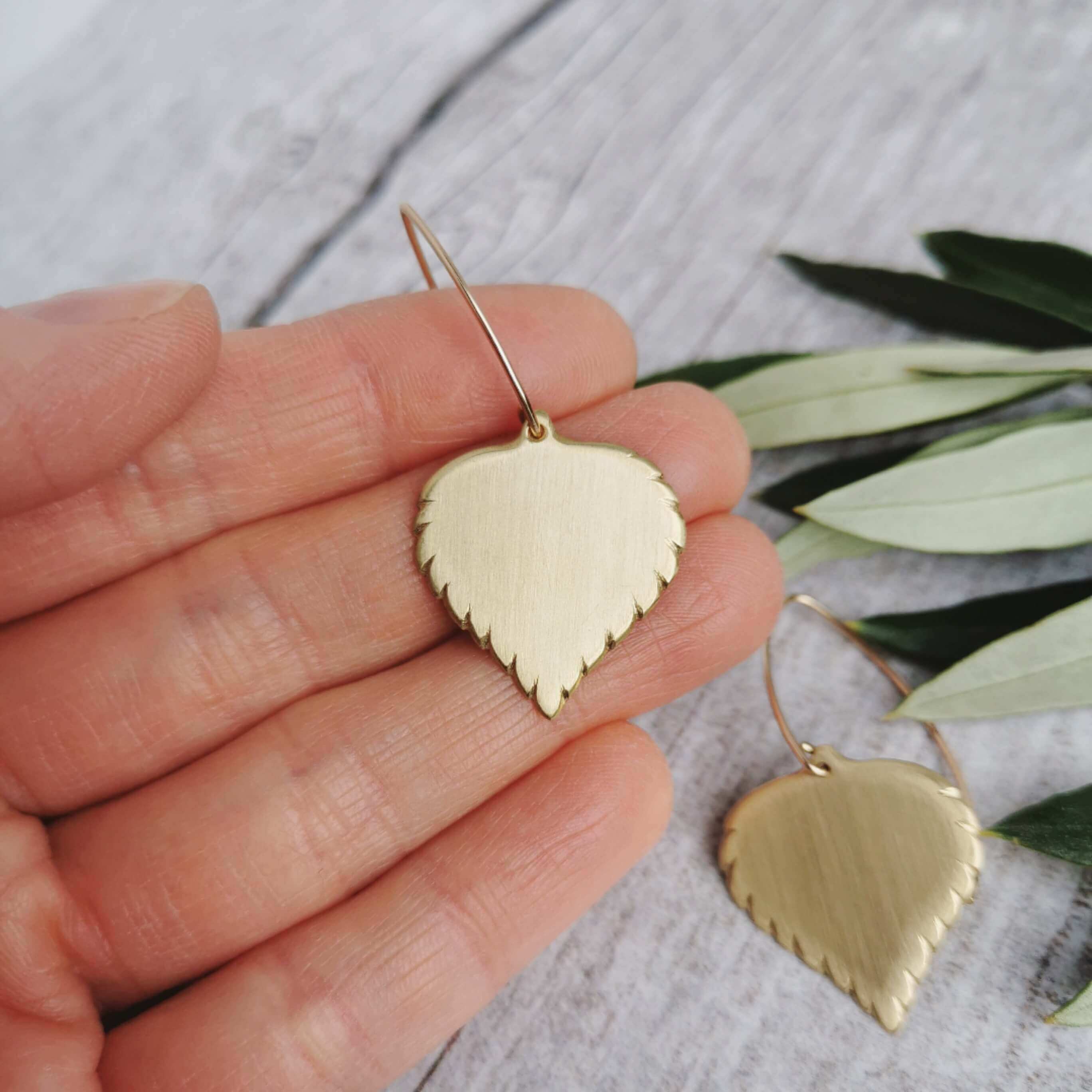 Golden brass birch leaf-shaped earrings held in a hand with a light background