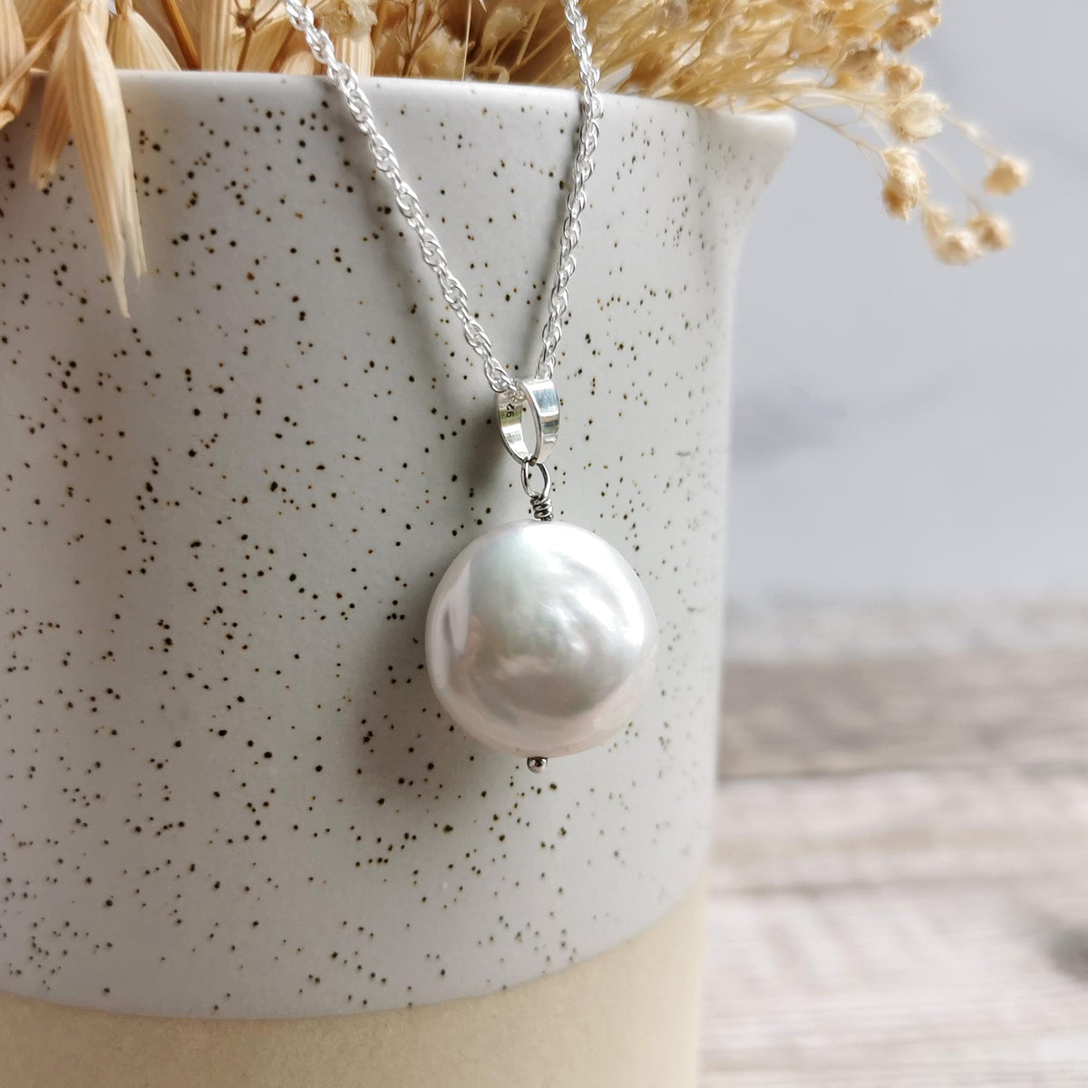 A silver chain statement necklace with a large, lustrous white coin pearl pendant, draped over a speckled ceramic vase with dried grasses.