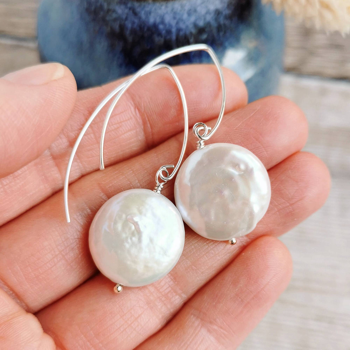 A pair of silver earrings with large, rippled ivory white coin pearls, held in a person's hand against a blurred blue ceramic background.