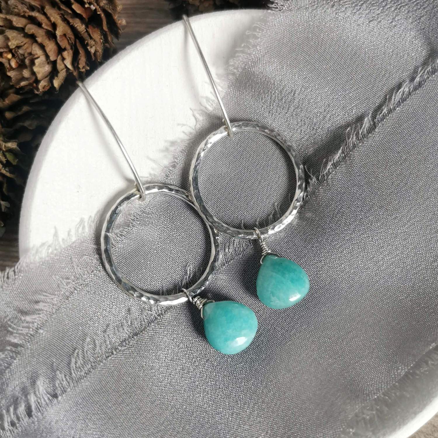 A pair of silver circle drop earrings with hammered texture and dangling amazonite teardrop charms, displayed on a textured gray fabric over a white circular surface.