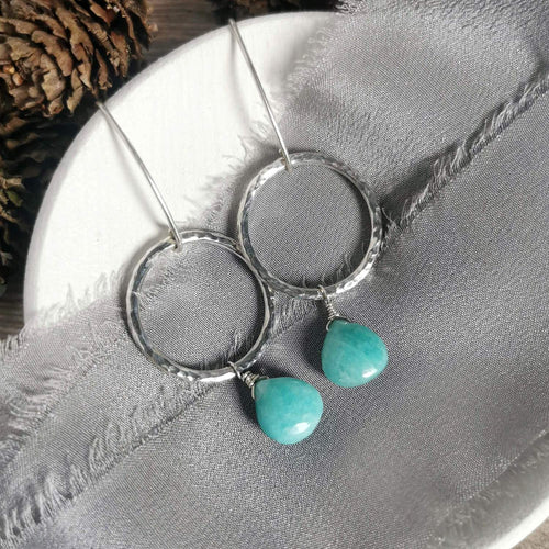 A pair of silver circle drop earrings with hammered texture and dangling amazonite teardrop charms, displayed on a textured gray fabric over a white circular surface.