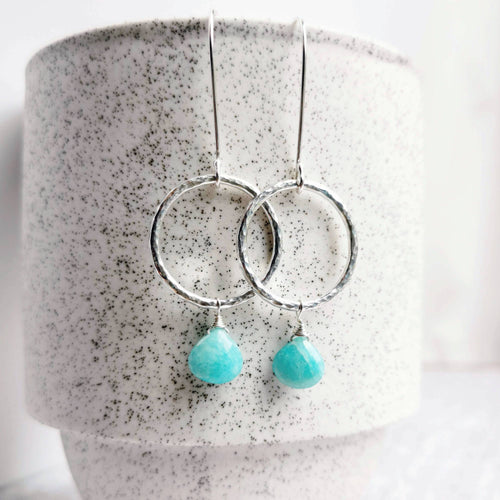 A pair of textured silver circle earrings with dangling turquoise coloured amazonite teardrop beads, displayed against a speckled white ceramic cup.