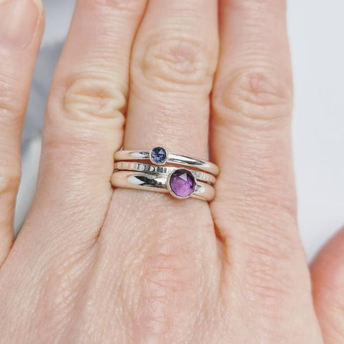 Three silver rings with purple and blue coloured gemstones - amethyst & iolite - on a hand against a neutral background