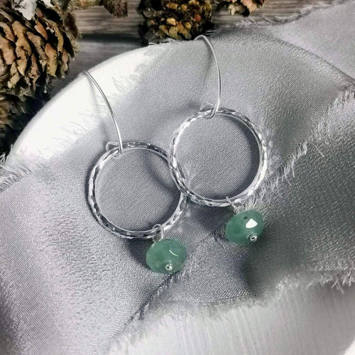 A pair of silver earrings with textured circles and dangling light green faceted aventurine gemstone beads, displayed on a textured gray fabric background.