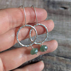 A pair of silver circle earrings with hammered edges and small green faceted gemstone bead drops, held in a person's hand against a rustic wooden background.