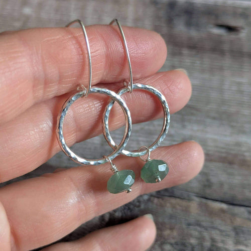 A pair of silver circle earrings with hammered edges and small green faceted gemstone bead drops, held in a person's hand against a rustic wooden background.