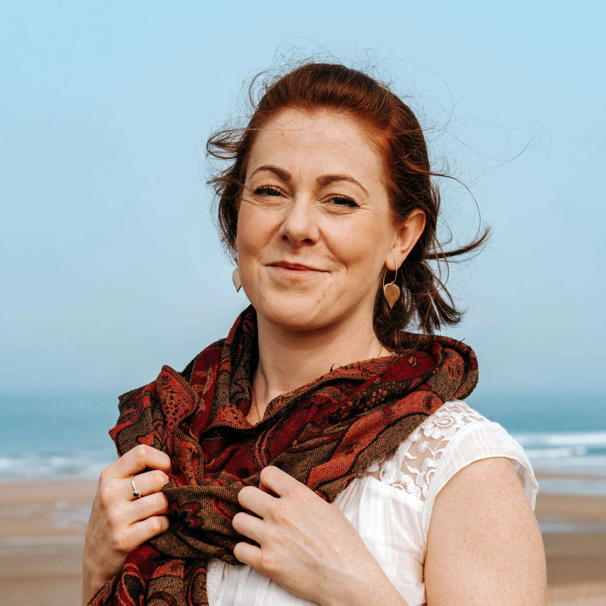 Woman wearing brass birch leaf shaped handmade earrings holding a patterned scarf on a cornish beach with a clear blue sky.