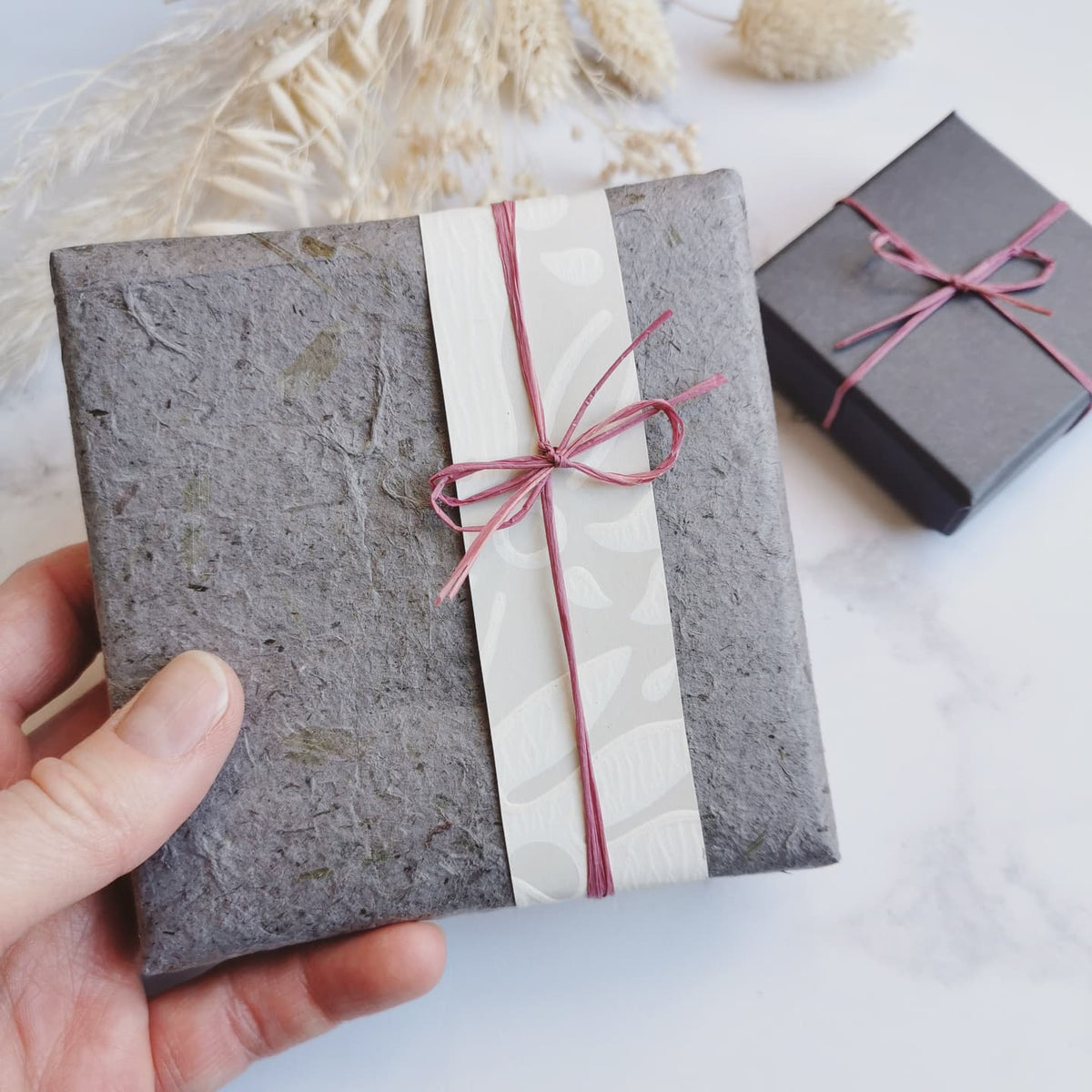 A hand holds a square gift box wrapped in textured grey paper with a white patterned paper band and a dusty pink ribbon tied in a bow. A second dark gray box sits in the background on a light marble surface with dried grass decor.
