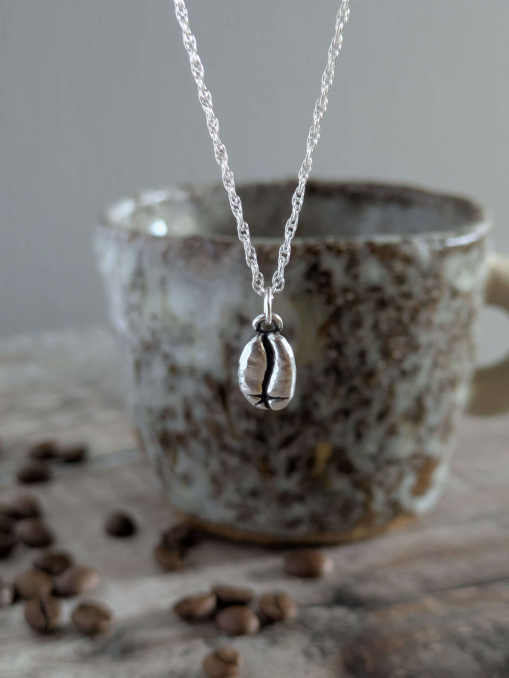 Silver charm necklace with a silver coffee bean pendant in front of a ceramic mug with coffee beans on a wooden surface.