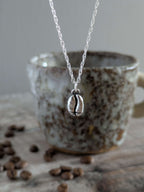 Silver charm necklace with a silver coffee bean pendant in front of a ceramic mug with coffee beans on a wooden surface.