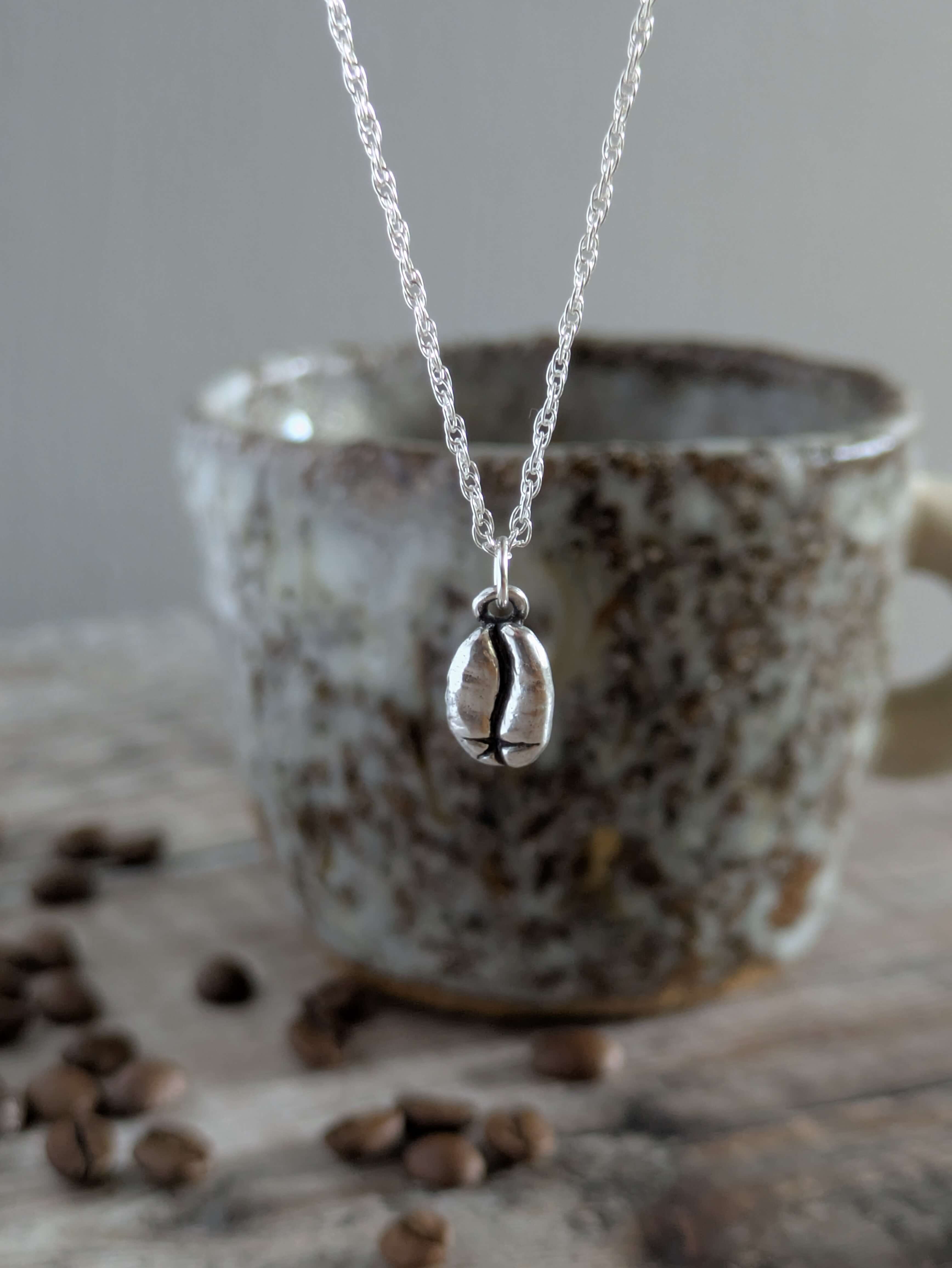 Silver charm necklace with a silver coffee bean pendant in front of a ceramic mug with coffee beans on a wooden surface.