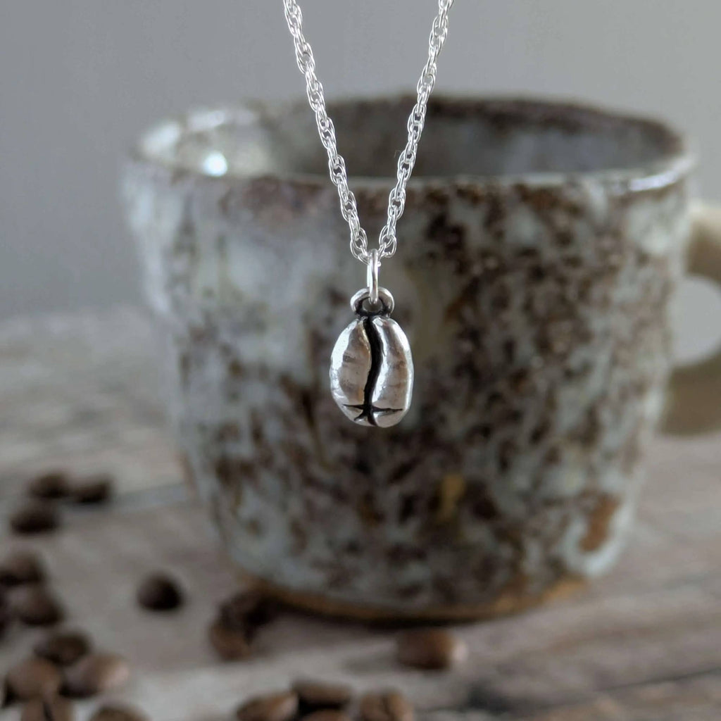Silver necklace with a coffee bean charm in front of a ceramic mug with coffee beans on a wooden surface.