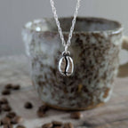 Silver necklace with a coffee bean charm in front of a ceramic mug with coffee beans on a wooden surface.