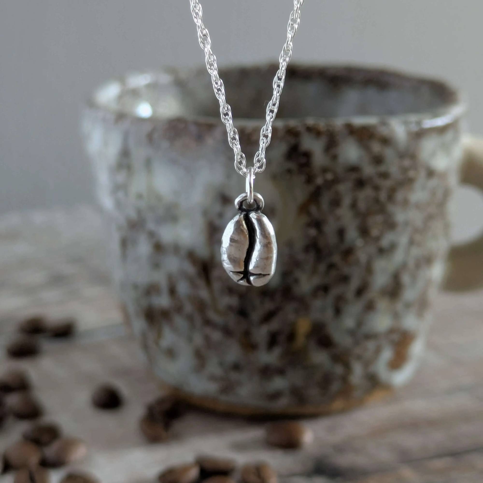 Silver necklace with a coffee bean charm in front of a ceramic mug with coffee beans on a wooden surface.