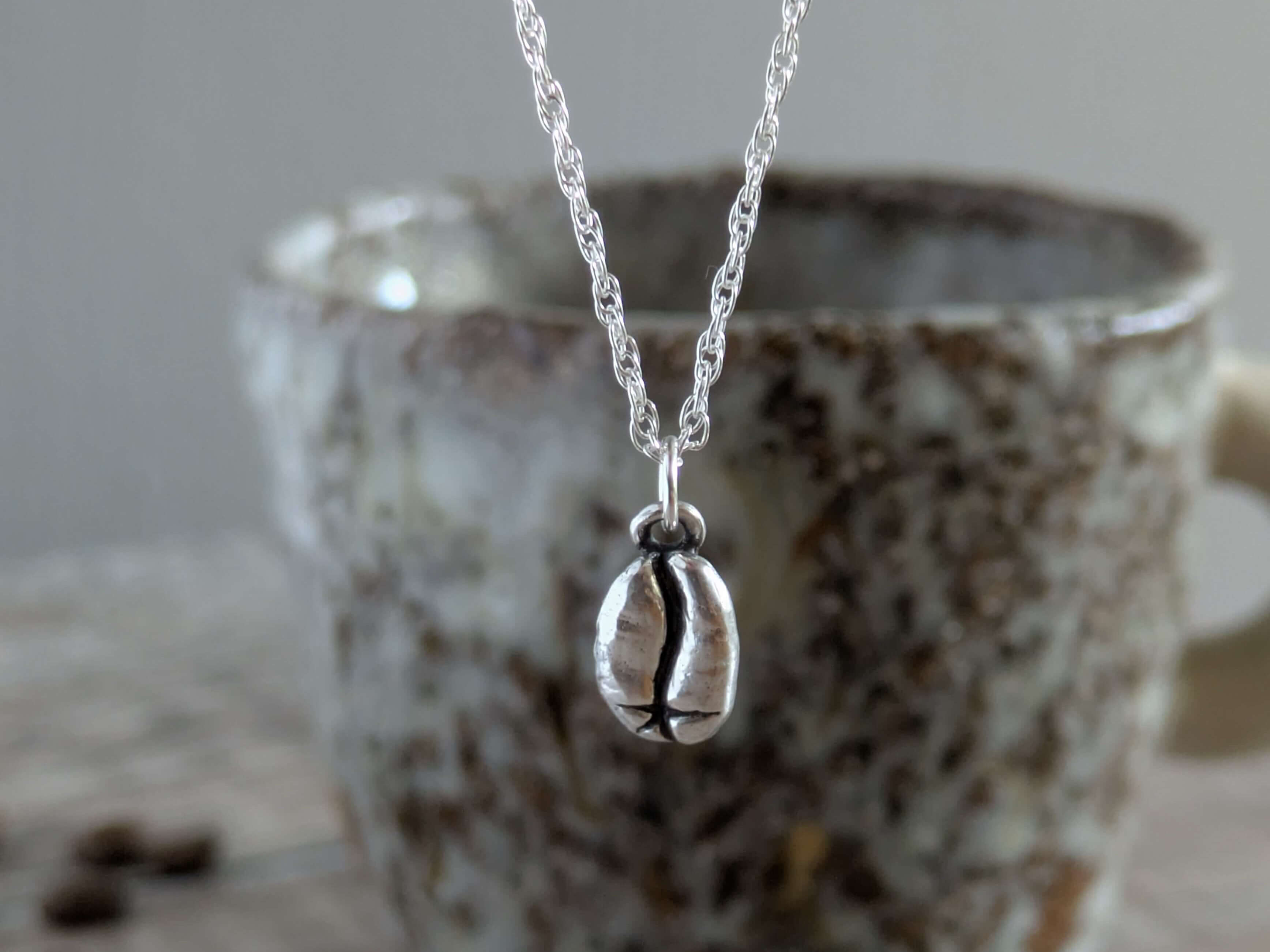 handmade silver necklace with a silver coffee bean pendant in front of a ceramic mug