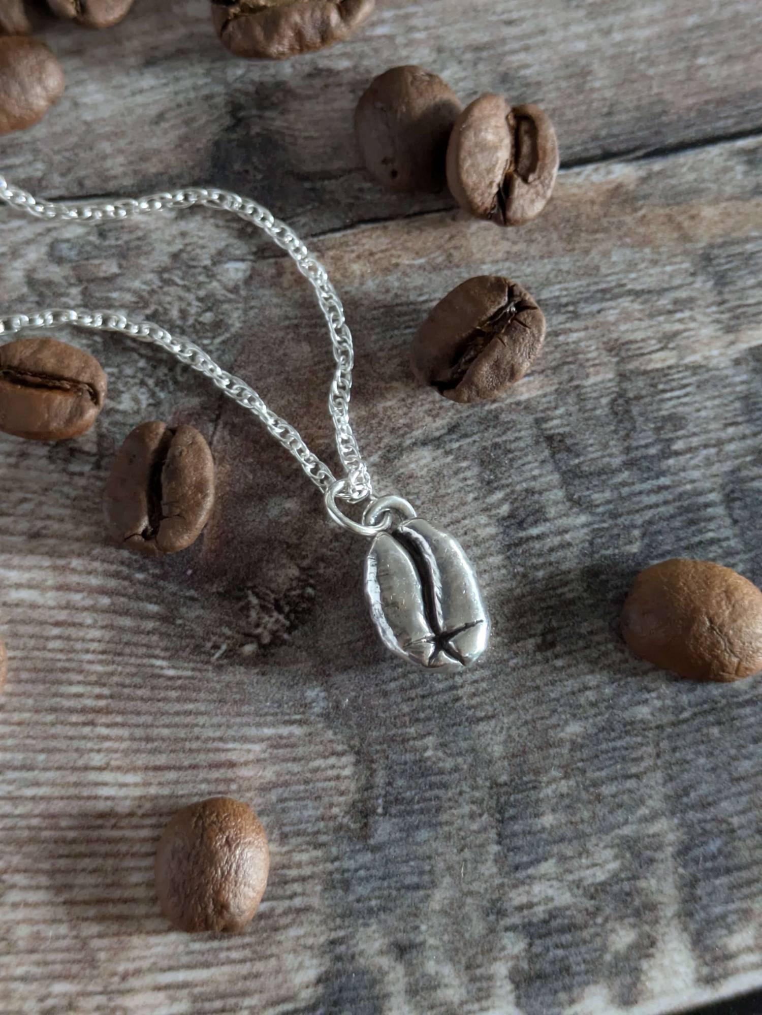 Silver necklace with a coffee bean charm on a wooden surface with coffee beans.