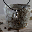 Necklace with a coffee bean pendant on a leather cord in front of a ceramic mug with coffee beans on a wooden surface.