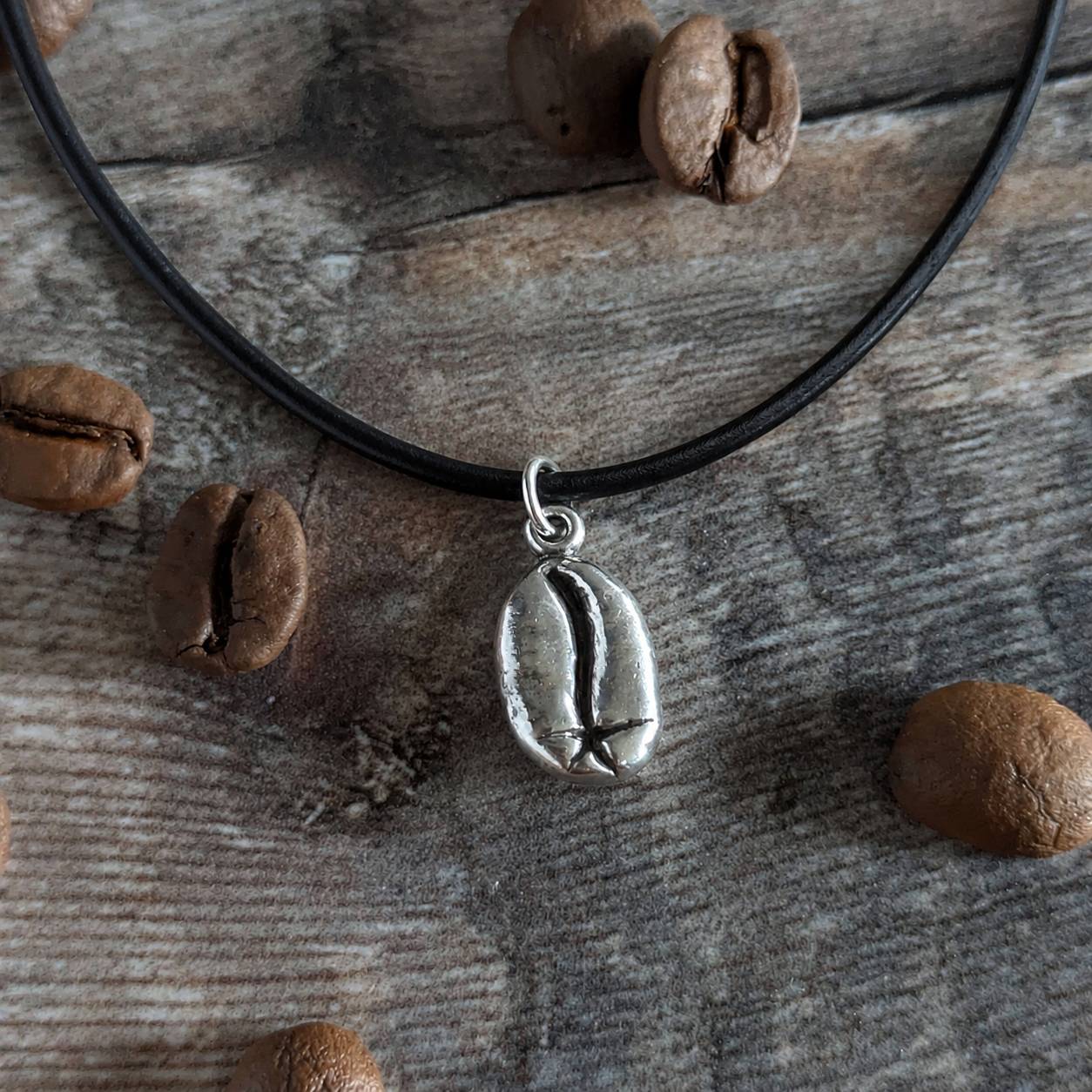 Necklace with a silver coffee bean pendant on a leather cord, rested on a wooden surface with coffee beans.