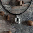 Necklace with a silver coffee bean pendant on a leather cord, rested on a wooden surface with coffee beans.