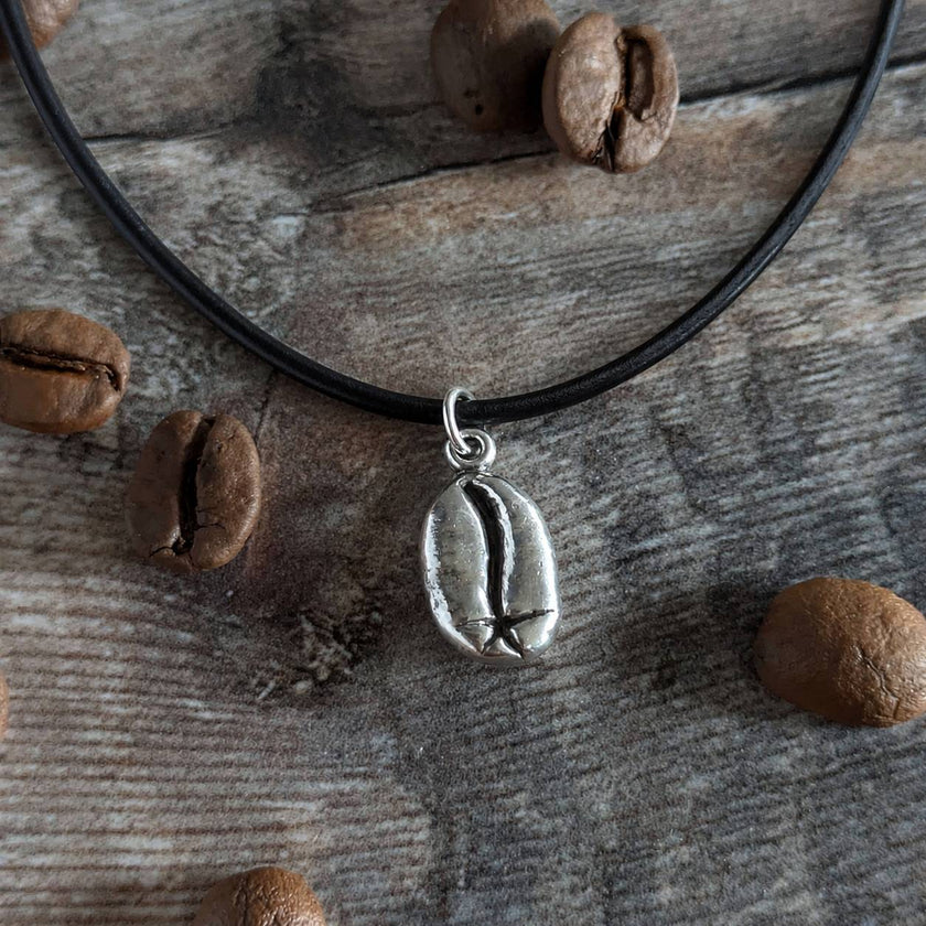 Necklace with a silver coffee bean pendant on a leather cord, rested on a wooden surface with coffee beans.