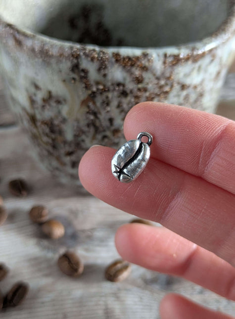 Silver coffee bean-shaped silver charm held between fingers with a cup and beans in the background