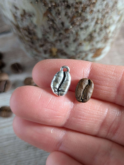 Silver coffee bean charm and a real coffee bean on a finger with a blurred coffee cup and coffee beans in the background.