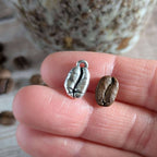 Silver coffee bean charm and a real coffee bean on a hand with blurred coffee beans in the background.