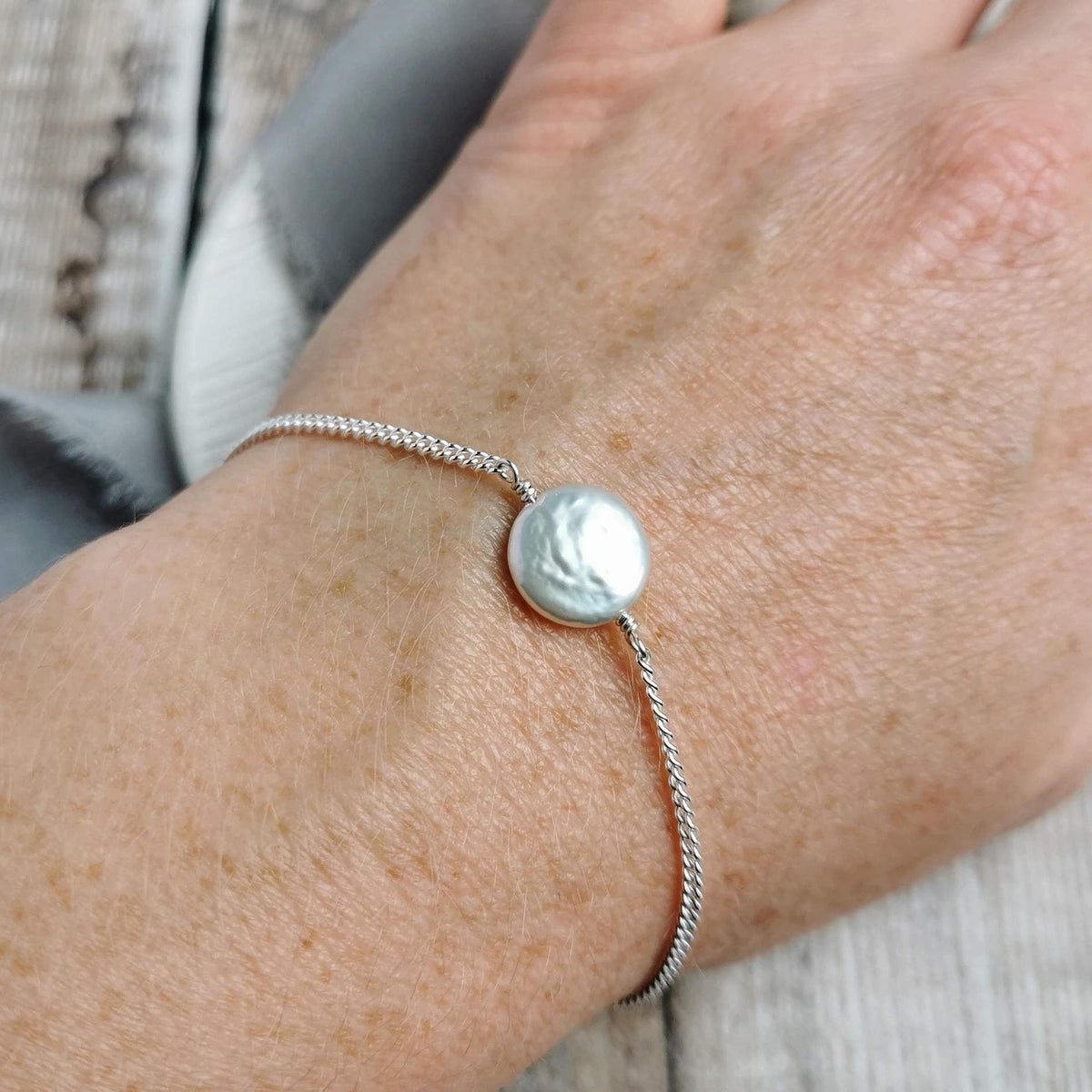 A close-up of a person's wrist wearing a delicate silver chain bracelet with a single round, coin shaped lustrous ivory freshwater pearl.
