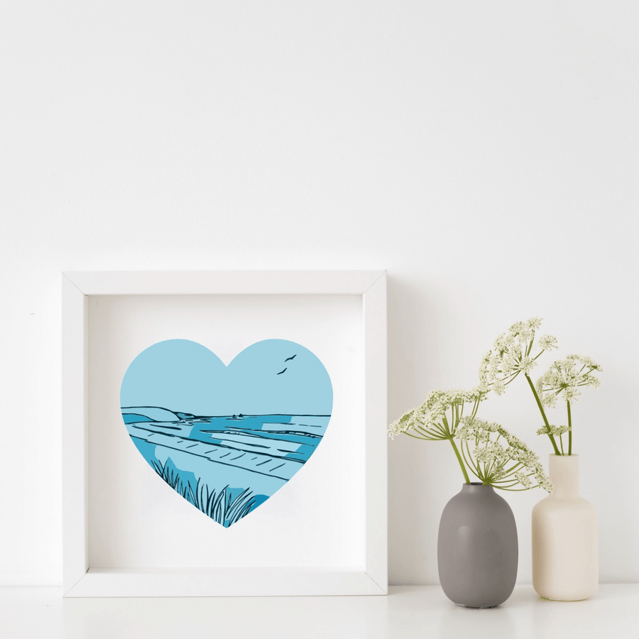 Framed artwork with a blue heart design of a beach scene, placed on a white surface with two small vases.