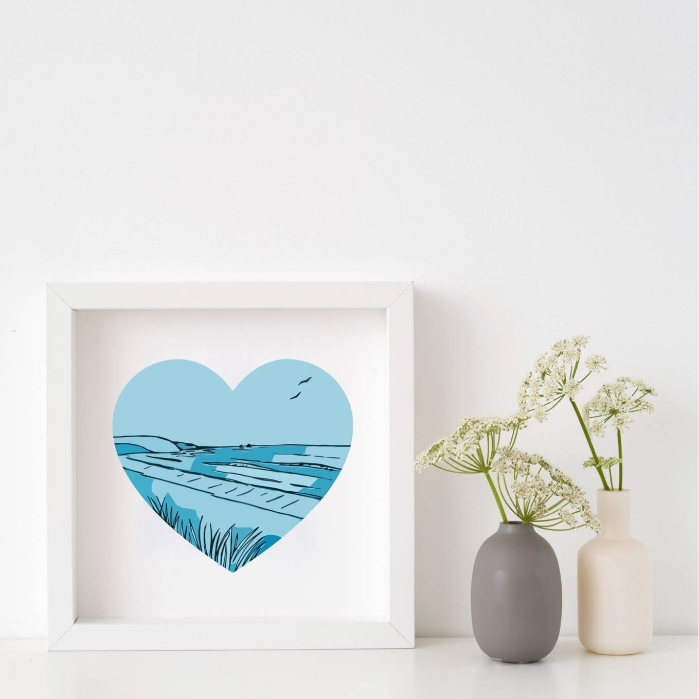 Framed artwork of a heart-shaped landscape in blues, placed on a white surface with handmade ceramic vases.