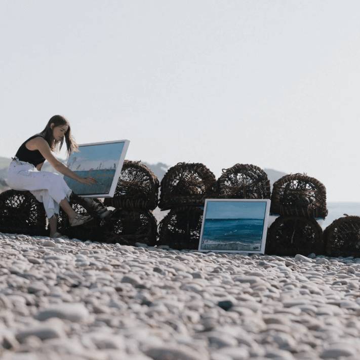 Woman sitting on a pebbly beach with two framed artworks, one of which she is still painting.