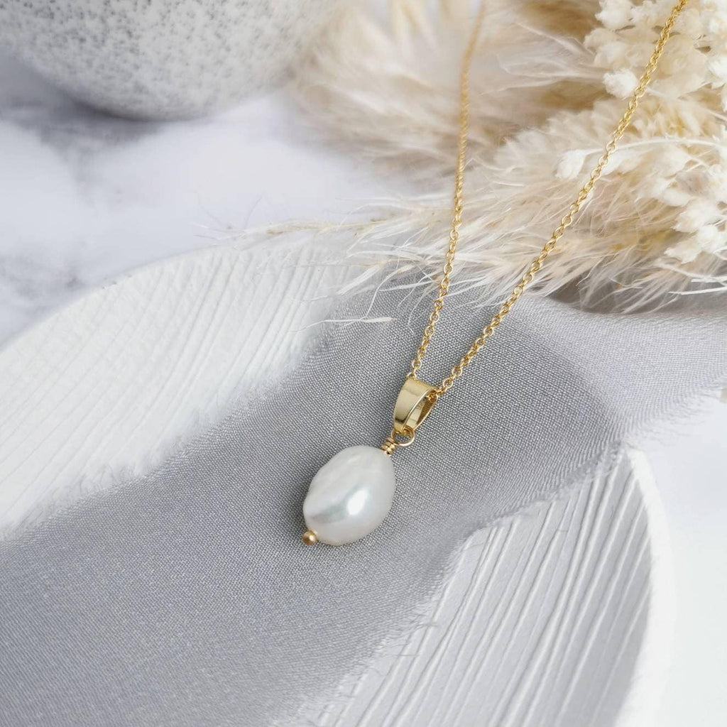 A silver or 14k gold-filled pendant necklace with a single baroque pearl centerpiece, presented on a grey fabric surface with dried flowers.