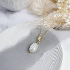 A silver or 14k gold-filled pendant necklace with a single baroque pearl centerpiece, presented on a grey fabric surface with dried flowers.