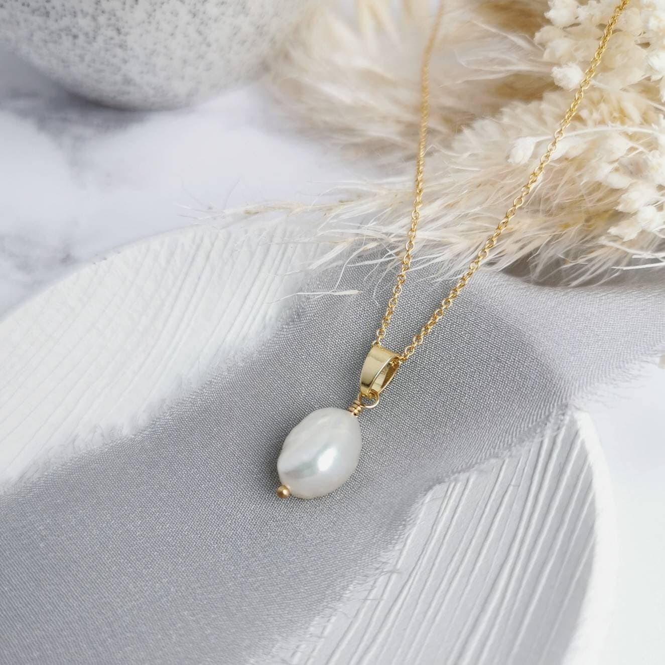 A silver or 14k gold-filled pendant necklace with a single baroque pearl centerpiece, presented on a grey fabric surface with dried flowers.