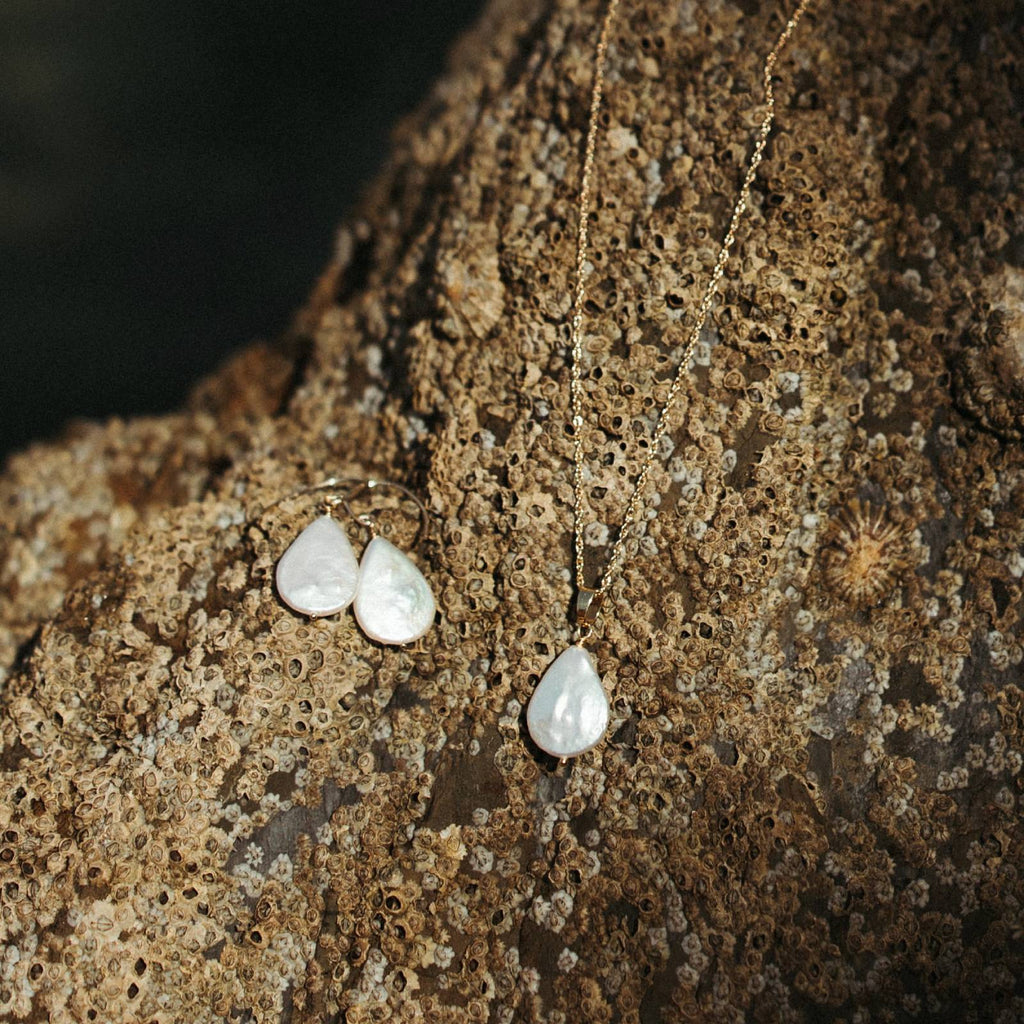 A set of 14k gold-filled necklace and matching drop earrings with ivory teardrop pearls, displayed on a textured brown rock covered in barnacles in cornwall, UK.