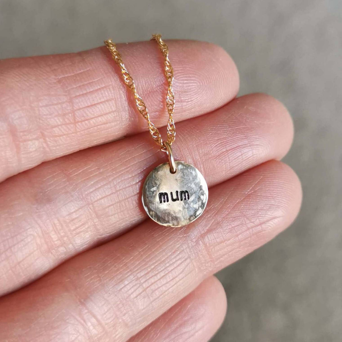 A handmade gold birthstone necklace with a round, hammered disc pendant stamped with 'mum' hidden on the back, held on a person's hand against a soft gray background.