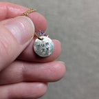 A close-up of a hand holding a small gold chain necklace with a round gold disc pendant stamped with 'mum xx' and small pink and purple tourmaline, amethyst and opal gemstones, against a neutral gray background.