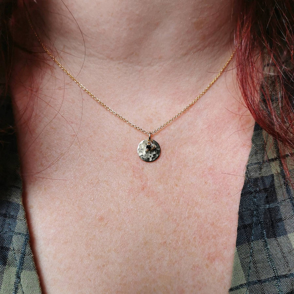 A close-up of a person's neck and chest wearing a delicate gold chain necklace with a small, textured circular pendant with two birthstones of sapphire for september and topaz for november.