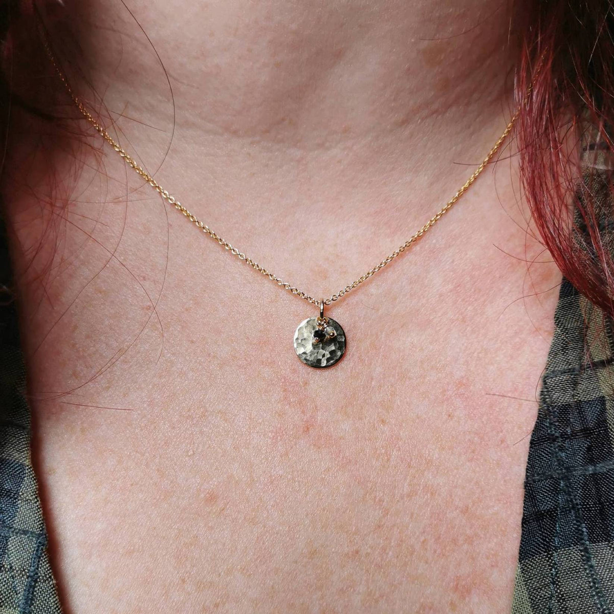 A close-up of a person's neck and chest wearing a delicate gold chain birthstone necklace with a small round, hammered-texture pendant with sapphire and topaz charms for september and october birth months