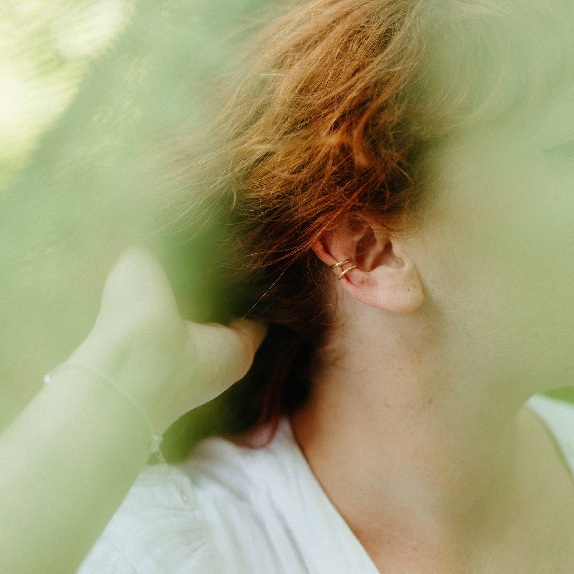 A close-up lifestyle shot of a person with short, wavy red hair wearing two delicate gold ear cuffs. The image has a soft, dreamy aesthetic looking through the ferns that are blurred 