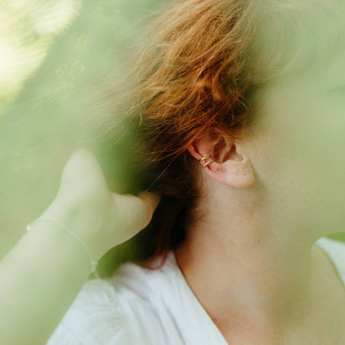 A close-up lifestyle shot of a person with short, wavy red hair wearing two delicate gold ear cuffs. The image has a soft, dreamy aesthetic looking through the ferns that are blurred 