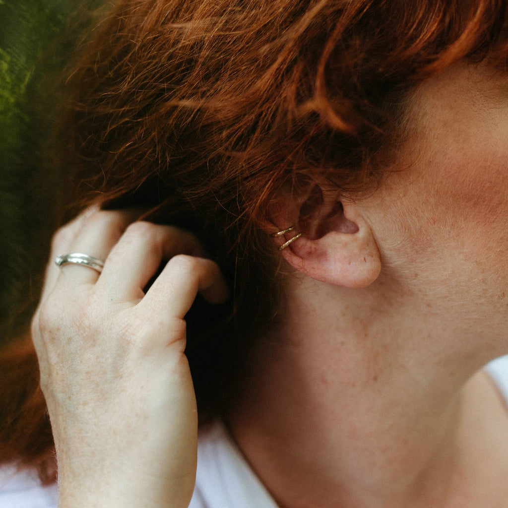 A close-up of a person's ear wearing two delicate gold ear cuffs, with red hair and a hand touching the hair in the background.