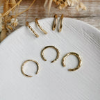 Set of gold ear cuffs in a variety of thicknesses and textures on a white surface with dried flowers in the background