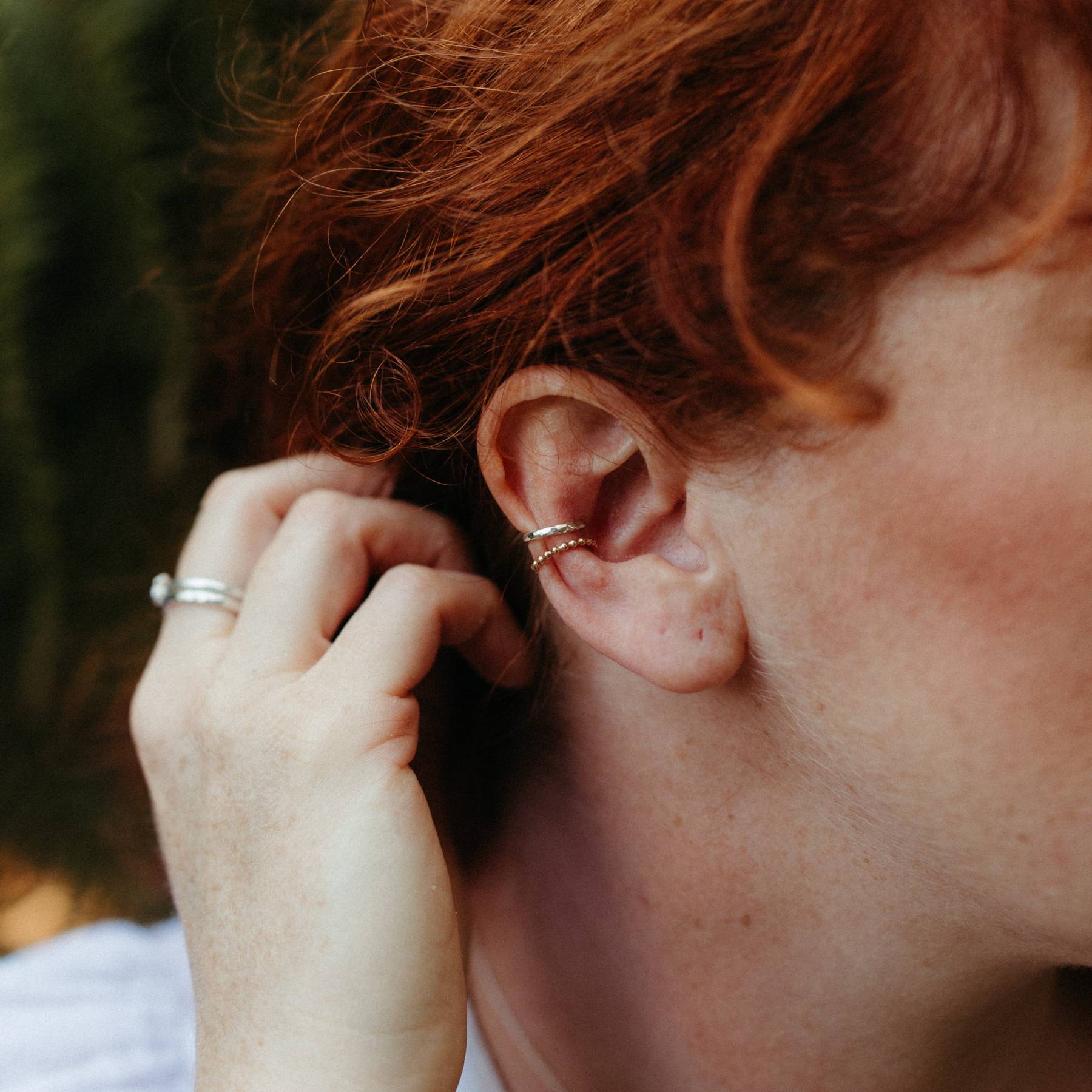woman with wavy red hair wearing a gold beaded decorative ear cuff and a hammered silver ear cuff and tucking her hair behind her ears