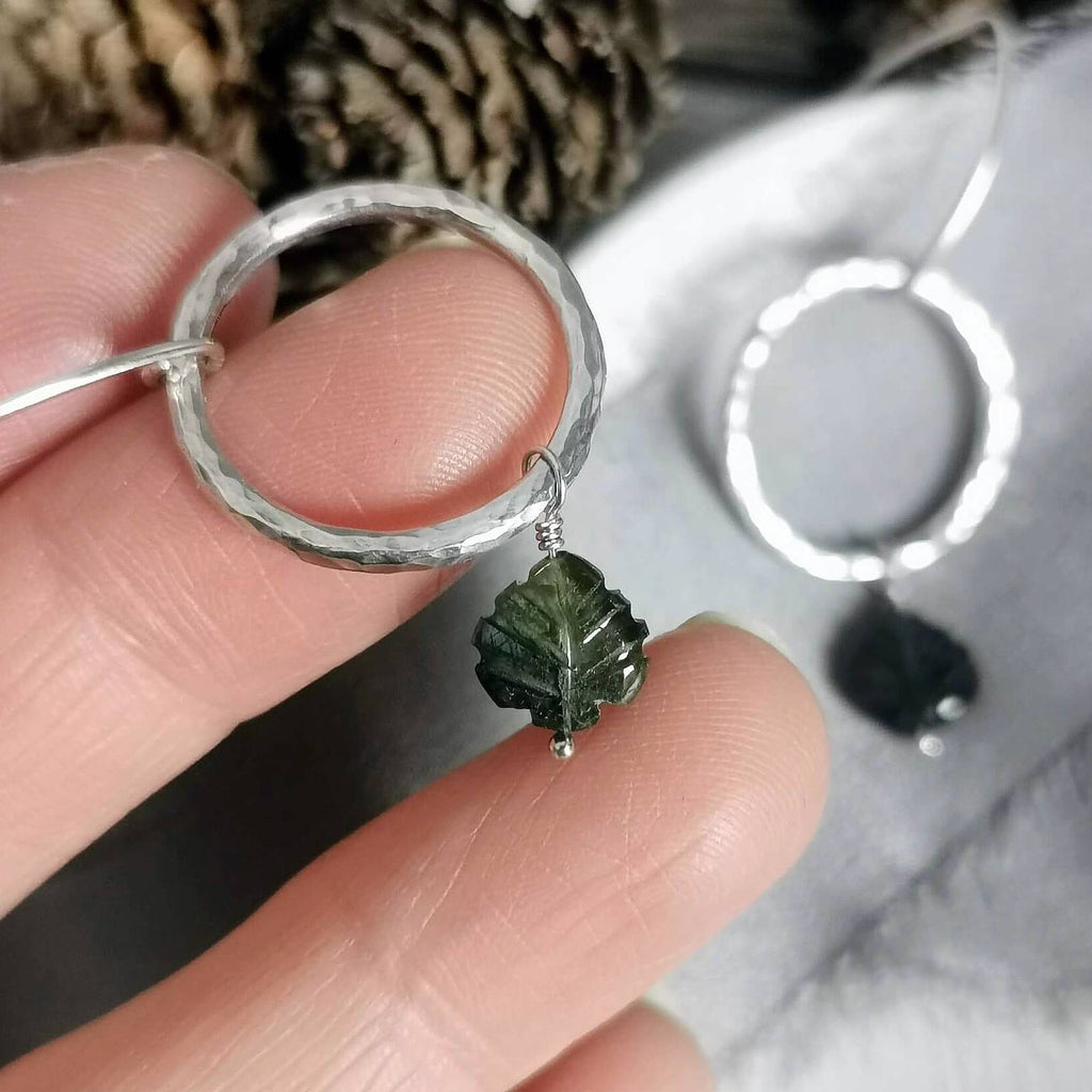 A close-up of a hand holding a hammered silver circle drop earring with a small carved green tourmaline leaf charm, with a second matching earring resting on a soft gray surface in the background.
